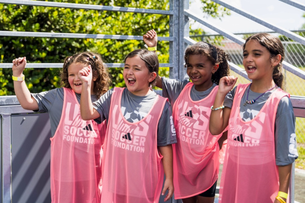 Four young girls stand side by side, wearing gray US Soccer Foundation t-shirts and pink pinnies. Each child raises their right fist, cheering.